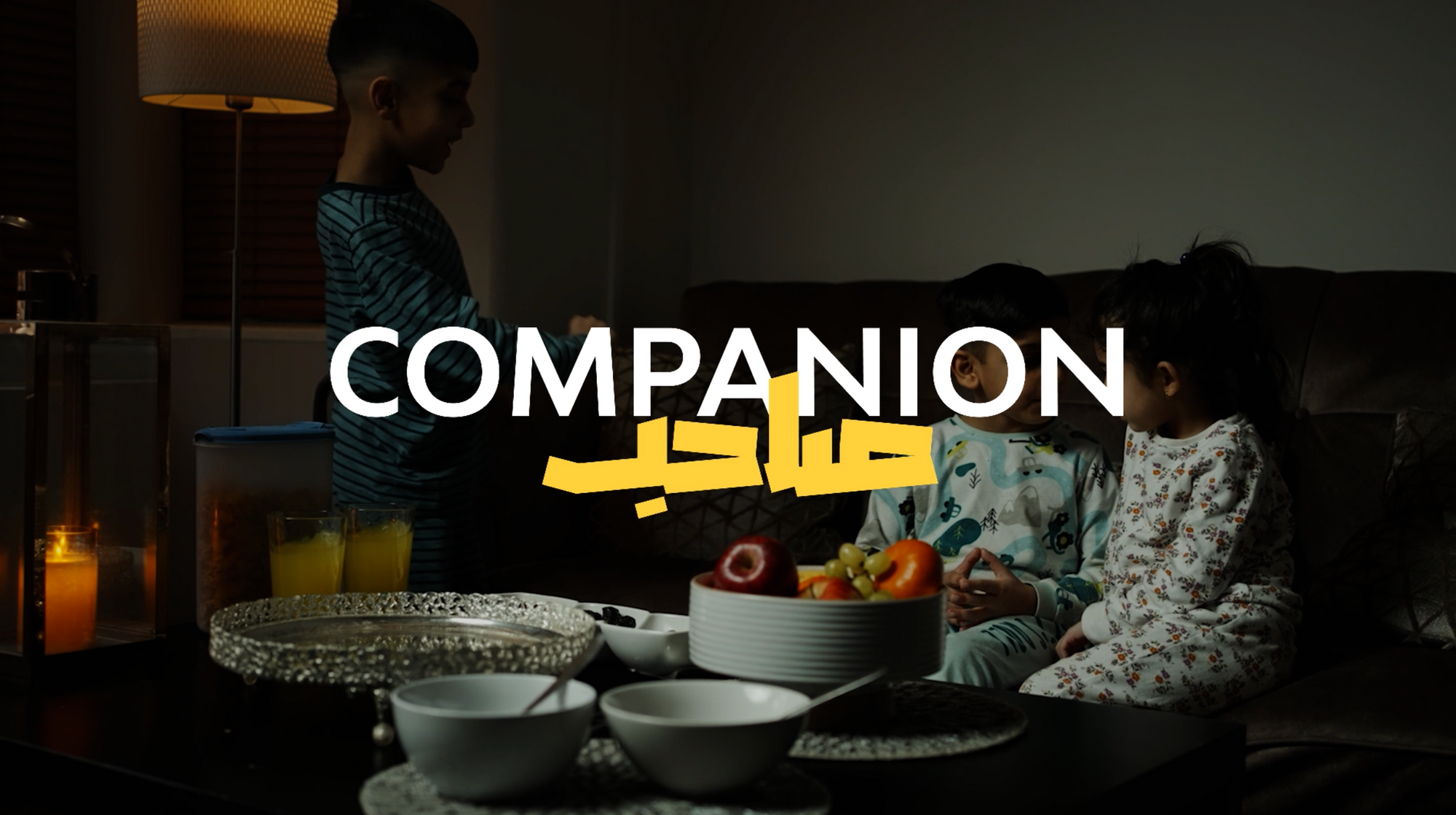 A photo of children around the coffee table in their living room. The text laid over says Companion in English and Arabic